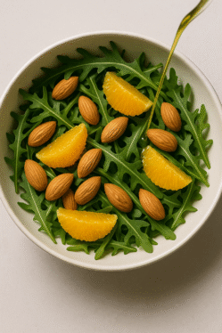 Insalata di rucola, mandorle sgusciate bio e spicchi d’arancia con condimento di olio EVO siciliano a crudo, ripresa dall’alto.